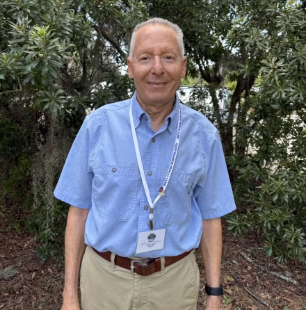 An older man wearing a light blue shirt, khaki pants, and a lanyard with a badge stands smiling in front of green trees and foliage outdoors.